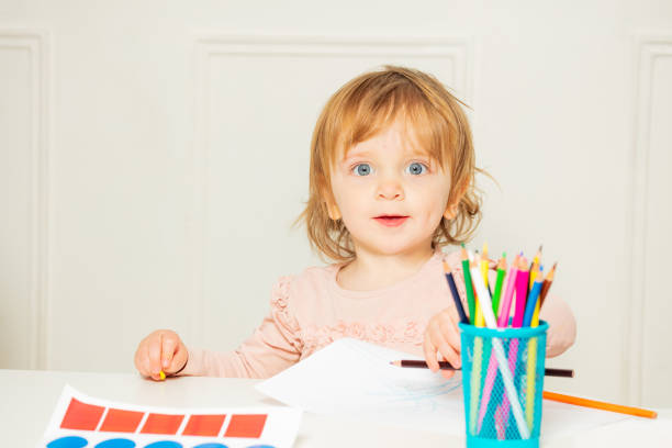 Little beautiful girl sit by the table and draw picture with pencil smiling look at the camera
