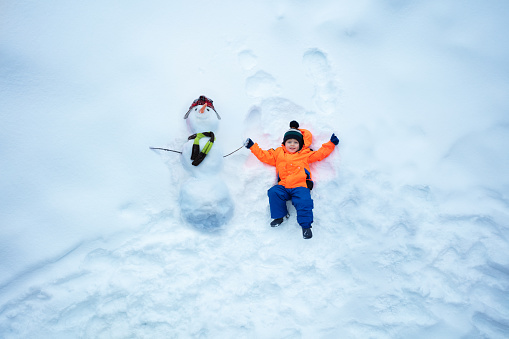Little boy in sport winter orange coat lay holding hand of snowman top view from above