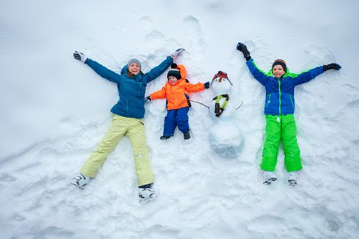 Family, mother and two boys lay in the snow with snowman view from top above, happy waving hands smiling during winter vacation