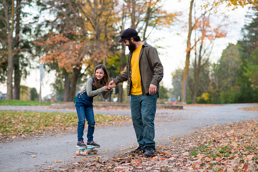 Little girl in jean overalls is holding her father's hand as she is riding a small skateboard for the first time and learning to balance. She is riding on the pavement of a public park, surrounded by trees and smiling while looking forward.