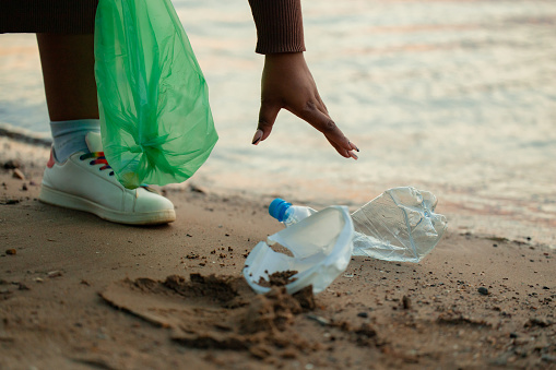 Cropped photo of African woman picking up spilled trash garbage from sand on beach in green plastic bag. Womans hand cleaning up used plastic bottles. Ecology, environmental conservation, pollution.