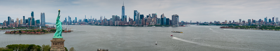 Panoramic drone shot of New York Harbor on an overcast day, looking past the Statue of Liberty and Ellis Island towards Jersey City, Manhattan, and Brooklyn.