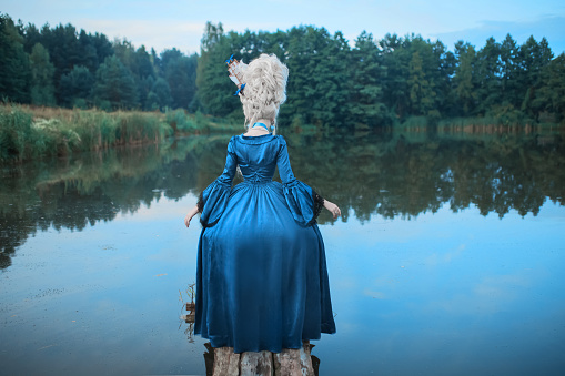 Renaissance princess with blonde hair on lake background. Beauty makeup. Fairytale rococo queen with ship in hairstyle on nature. Model in blue dress. Woman with historical hair style on bridge