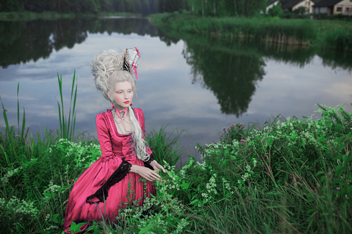 Renaissance princess with blonde hair on lake background. Beauty makeup. Fairytale rococo queen with ship in hairstyle on nature. Model in pink dress. Woman with historical hair style sit in grass.