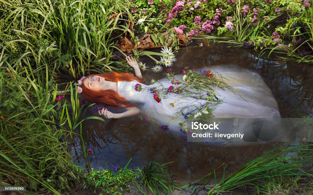 Fairy redhead princess in white dress in stream. Girl with long hair with flowers in pond. Renaissance painting. Ophelia. Summer vegetation in nature Ophelia - Fictional Character Stock Photo