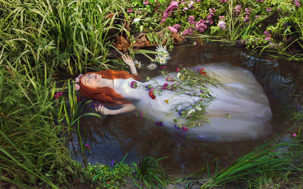 Fairy redhead princess in white dress in stream. Girl with long hair with flowers in pond. Renaissance painting. Ophelia. Summer vegetation in nature stock photo