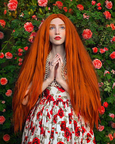 Redhead fabulous woman. Long straight hair. Pale skin. Girl with beads and flower dress on rose background. Beautiful model with red lips. Renaissance outfit. Hairstyle. Jewelry and bijouterie