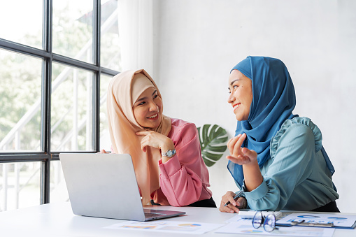 Young pretty Asian muslim female business worker discussing with friends sitting together, working with laptop at desk.