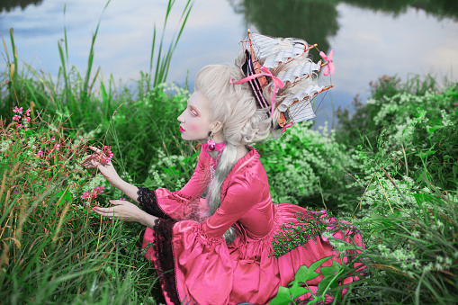 Renaissance princess with blonde hair on lake background. Beauty makeup. Fairytale rococo queen with ship in hairstyle on nature. Model in pink dress. Woman with historical hair style sit in grass.