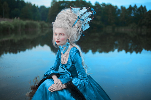 Renaissance princess with blonde hair on lake background. Beauty makeup. Fairytale rococo queen with ship in hairstyle on nature. Model in blue dress. Woman with historical hair style on bridge