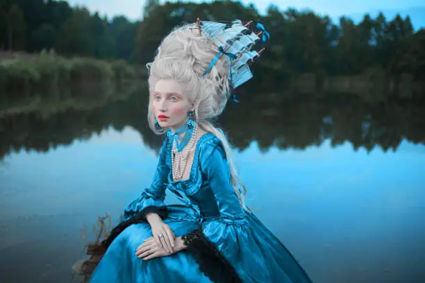 Photo of Renaissance princess with blonde hair on lake background. Beauty makeup. Fairytale rococo queen with ship in hairstyle on nature. Model in blue dress. Woman with historical hair style on bridge