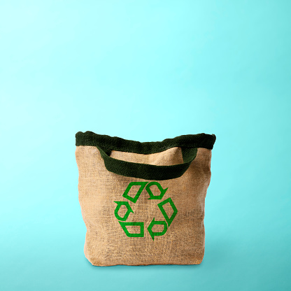 Burlap tote bag with recycling symbol on light blue background.\nConcept of environmental and recycling.