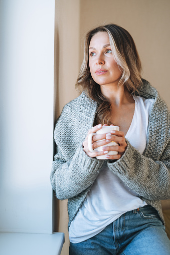Young beautiful woman forty year with blonde long curly hair in cozy knitted grey sweater with cup of tea in hands in bright interior at the home