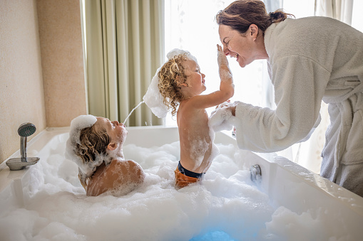 Mother and her boys having fun in the bathroom during bath time, making soap foam together in hot tub bath.