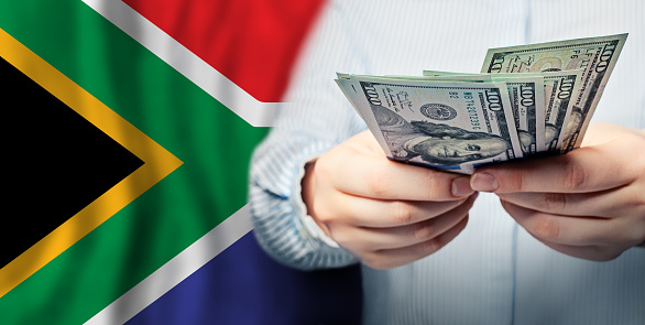 Hands holds American 100 dollars banknote and flag of South Africa closeup