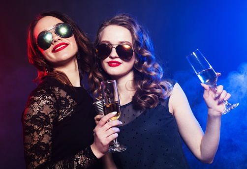 Lifestyle, friendship and party concept: two women in black dresses drinking wine and dancing over black background