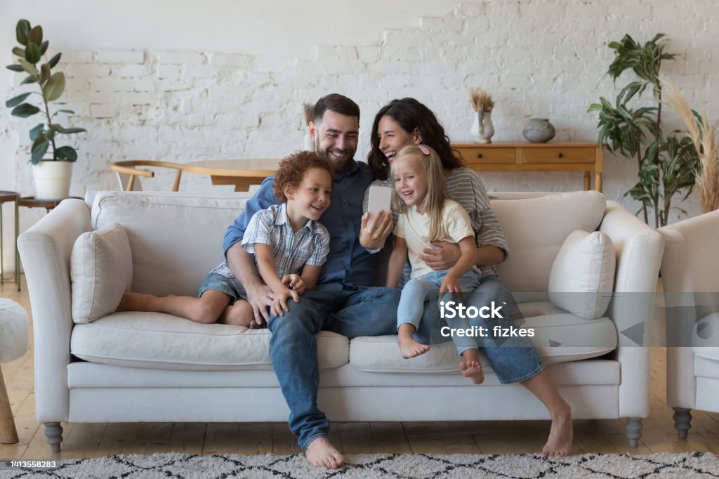 Happy parents, little son and daughter talking on video call, Happy excited couple of parents, little son and daughter kids talking on video call, taking selfie on cellphone, chatting, smiling, laughing, having fun, hugging on comfortable sofa at home Family Stock Photo