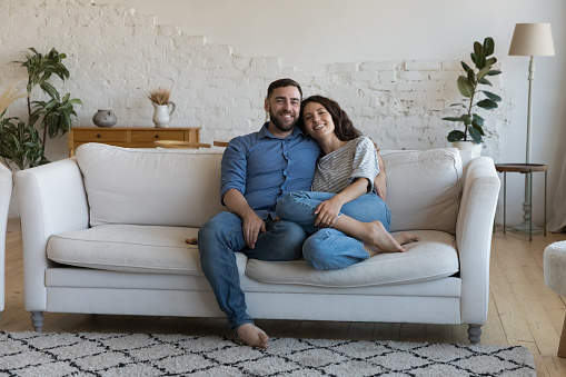 Happy young adult married couple home full length portrait. Millennial husband and wife hugging on cozy couch in new house, looking at camera, smiling, laughing. Relationship, love concept