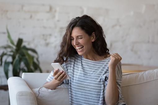 Excited smartphone user woman receiving good happy news, holding mobile phone, looking at screen, making winner hand, laughing, smiling, feeling joy, celebrating success, achieve