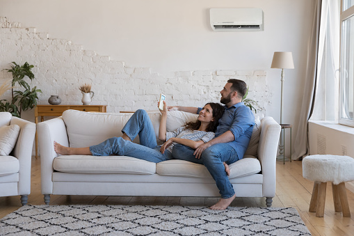 Happy millennial couple of homeowners enjoying cool conditioned air, resting on couch together, using remote control for AC, cooling domestic equipment start. Home appliance concept
