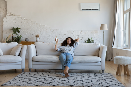 Happy pretty Latin woman enjoying cool conditioned air, relaxing on comfortable sofa, using remote control for turning AC on, starting cooling domestic equipment. Home appliance concept