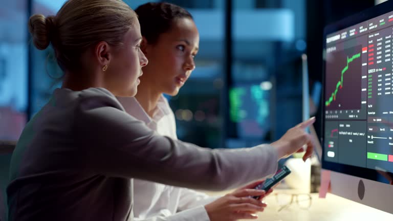 Finance business women talking and looking at data, stats and a graph on a computer screen while working late at night in an office. Female colleagues analysing a chart and trading stock online