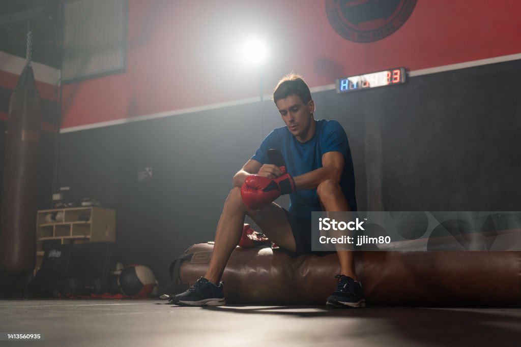 Young and handsome male boxer with red gloves sitting for a break after training in a gym Photo of a young and handsome boxer with muscular build body with red gloves sitting for a break after training in a gym 25-29 Years Stock Photo