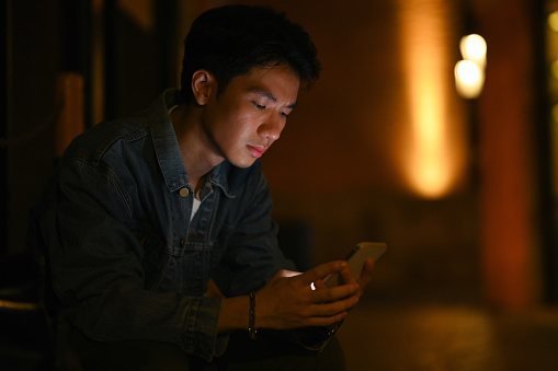Young hipster man using mobile phone, chatting with friends in social networks while sitting at evening city streets.