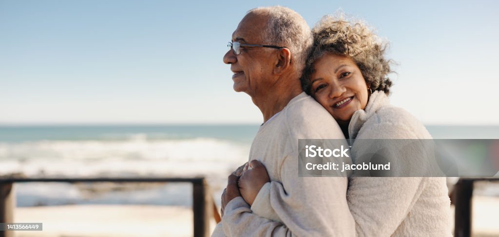 Romantic senior woman embracing her husband by the ocean Romantic senior woman smiling at the camera while embracing her husband by the ocean. Affectionate elderly couple enjoying spending some quality time together after retirement. Senior Couple Stock Photo