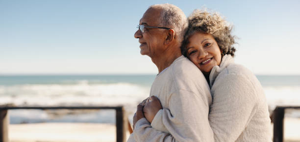 Romantic senior woman embracing her husband by the ocean Romantic senior woman smiling at the camera while embracing her husband by the ocean. Affectionate elderly couple enjoying spending some quality time together after retirement. senior lifestyle stock pictures, royalty-free photos & images