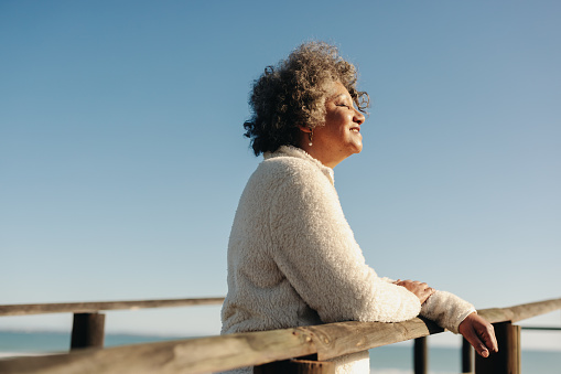 Enjoying the sun at the beach. Happy senior woman getting some sunlight while standing on a boardwalk next to the ocean. Cheerful elderly woman taking a refreshing holiday after retirement.