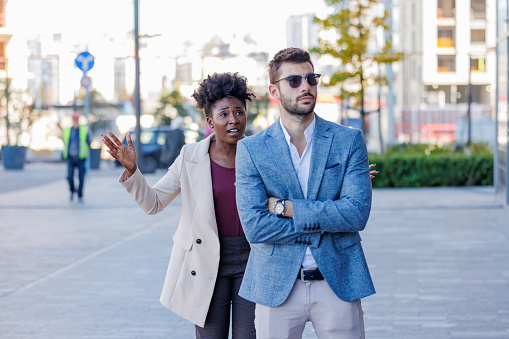 A young African American woman is standing with her arms raised in frustration and a negative facial expression, while her young Caucasian boyfriend ignores her with his back turned.