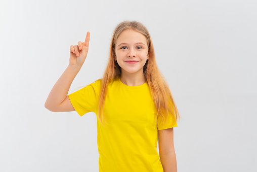 Happy cute teen girl pointing finger up, showing promo offer, way to store, banner or logo, standing in blank yellow tshirt over white background