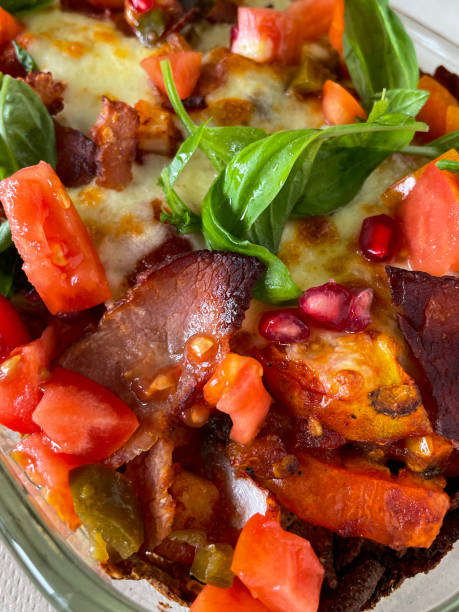 Stock photo showing close-up view of glass, ovenproof serving dish of homemade loaded nachos recipe. The cooking dish contains fresh, crunchy tortilla chips covered with tomato salsa and melted cheese and then topped with chopped tomatoes, bacon pieces and basil herb leaves. imagen en primer plano de nachos cargados cubiertos de salsa de tomate casera y queso mozzarella derretido, cubiertos con tomates picados, trozos de tocino, semillas de granada y albahaca, vidrio, plato para servir a prueba de horno, listo para comer, vist