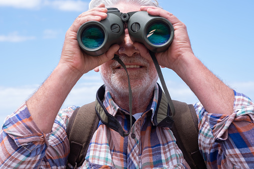 Close up on active senior man  in outdoors excursion using binoculars looking at camera, elderly caucasian male enjoying freedom and healthy lifestyle