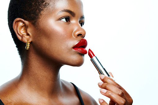 Black woman applying lipstick, makeup and beauty cosmetics to her face while posing against a white studio background. Closeup of one beautiful, serious and young female showing flawless skin