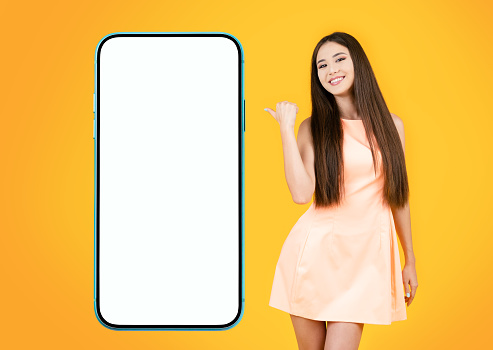 Beautiful asian woman showing her finger to blank empty cellphone on yellow background. She points to a profitable and interesting offer or application.