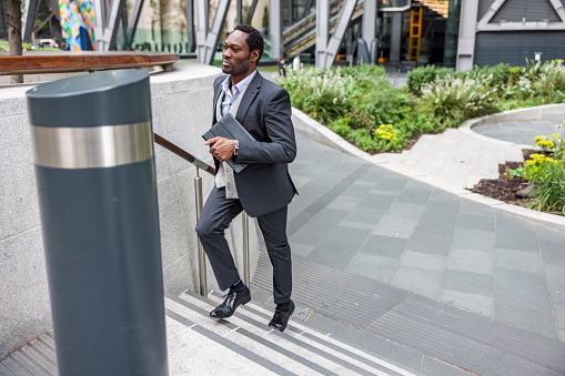 Serious Black male business strategist running upstairs. He is holding a digital tablet and wearing formal clothing.