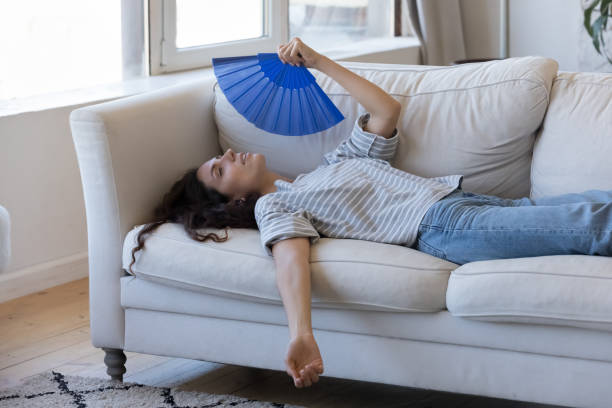 Exhausted young woman suffering from hot weather, overheat Exhausted young woman suffering from hot weather, overheat, too high air temperature at home, lying on sofa, waving paper handheld fan, cooling, feeling discomfort humidity stock pictures, royalty-free photos & images