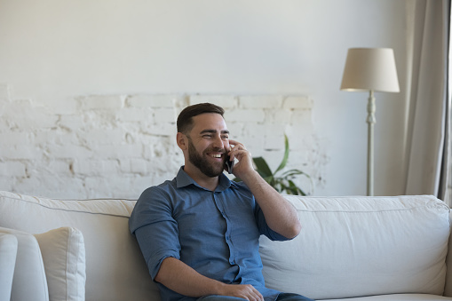 Cheerful millennial man talking on mobile phone at home, making telephone call, chatting, speaking, listening to good news, enjoying leisure, communication, laughing, smiling, resting on sofa