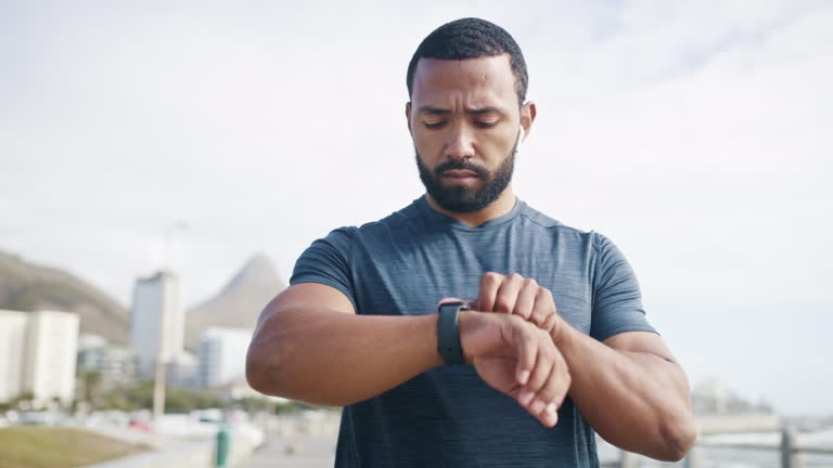 Athletic jogger tracking sprinting progress on smartwatch. Young man listening to music playlist through bluetooth airpods while running. Fit, strong athlete jogging on the promenade for stamina