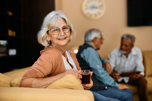 Smiling elderly woman drinking tea while enjoying residential care home and looking at camera.