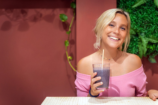 Young woman smiling at camera while enjoying drinking a delicious smoothie in a coffee shop. Healthy lifestyle concept.