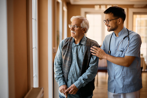 Male nurse assisting senior man who is standing by the window and looking through in at nursing home. Copy space.