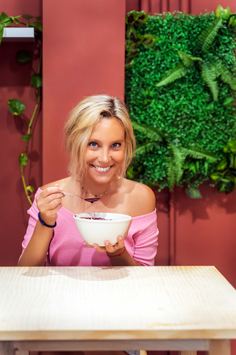 Woman smiling at camera while eating an acai smoothie bowl in a cafe. Healthy eating concept.