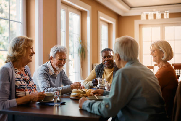Happy seniors talking while eating lunch at residential care home. Group of senior people enjoying in conversation during lunch at dining table at nursing home. senior lifestyle stock pictures, royalty-free photos & images