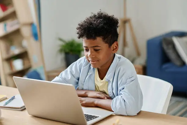 African schoolboy sitting at desk in his room and watching on screen of laptop, he studying online