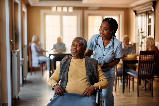 African American senior in wheelchair and young nurse looking through the window at residential care home.