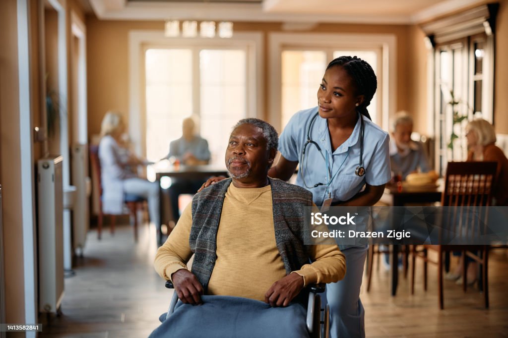 Black nurse and senior man in wheelchair looking through the window at nursing home. African American senior in wheelchair and young nurse looking through the window at residential care home. Nursing Home Stock Photo
