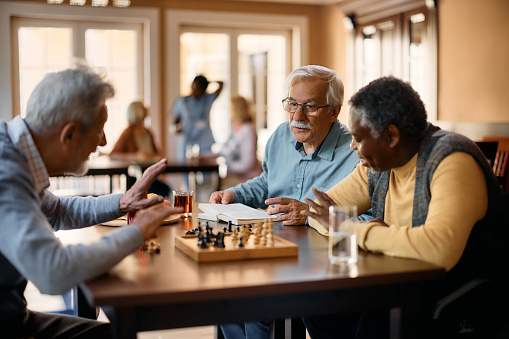 Mature man reading book and communicating with male friends while they are playing chess at retirement community.